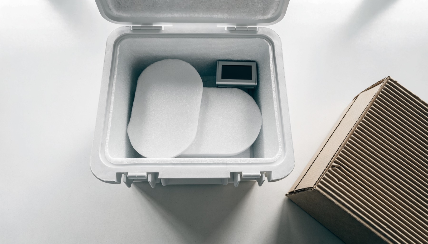 An opened white insulated shipping cooler on a light grey desk, lined with foam and holding two flat gel packs and a small electronic temperature data logger, beside a corrugated outer carton