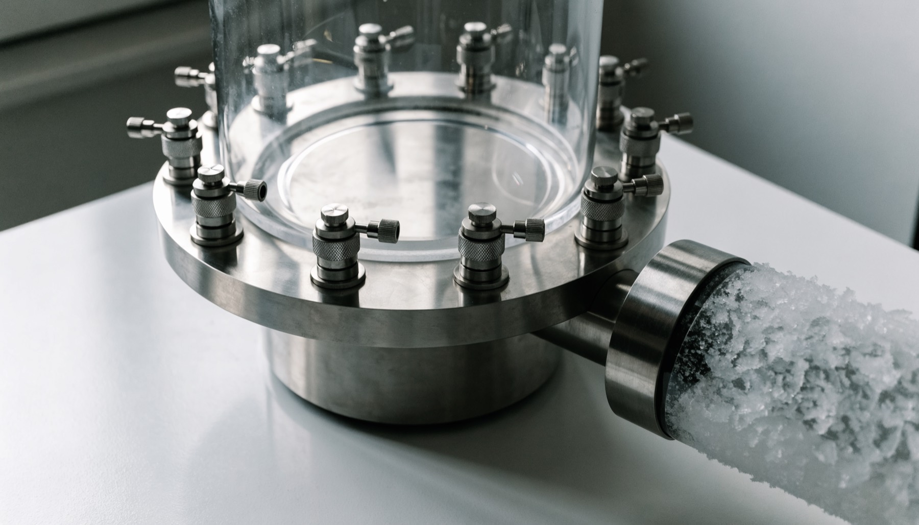 A benchtop freeze dryer: a clear cylindrical drying chamber on a stainless base with a radial manifold of ports and valves, and frost forming on the cold trap